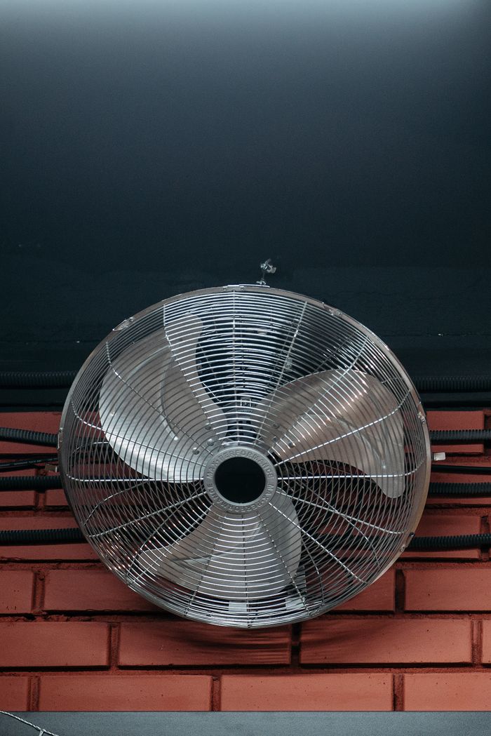 Close-up of an industrial ceiling fan with metal blades mounted on a brick wall indoors.